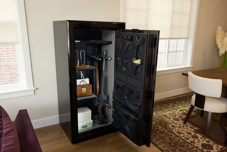 Open black Liberty gun safe in a home setting, showing interior shelving with stored items and a fabric-lined door panel.